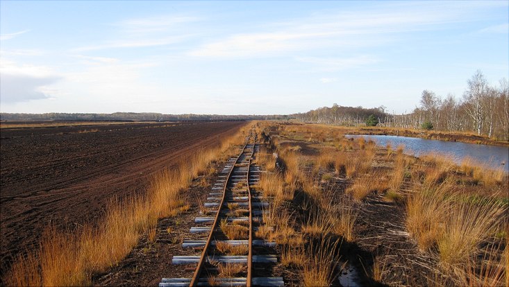 Weite Sicht über Himmelmoor, blauer Himmel, mittig alte Bahnschienen, links Feld, rechts Gewässer