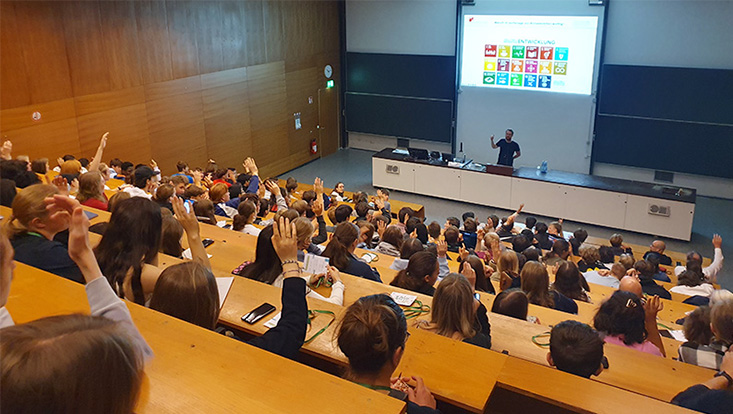 The picture shows students from the back of a lecture hall. In the front, Dr. Leonard Borchard is explaining something about the Sustainable Development Goals to the students.