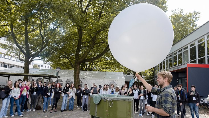 Professor und Schülerinnen auf dem Campus. Ein Experiment mit einem Wetterballon wird durchgeführt und erklärt.