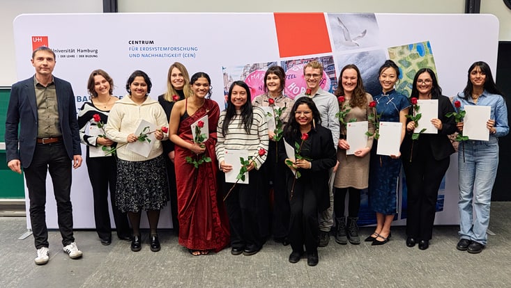 Prof. Christian Beer mit 12 Master-Absolvent:innen, die jeweils ihre Urkunde und eine Rose in den Händen halten.