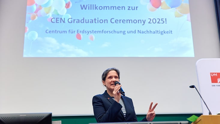 Prof. Johanna Baehr hält zwei Finger hoch, sie steht vor der Präsentation mit der Aufschrift "Willkommen zur CEN Graduation Ceremony 2025!"