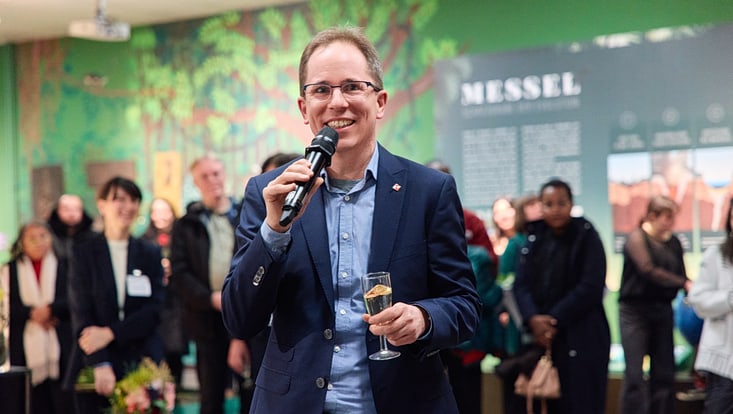 Prof. Grischa Perino spricht in ein Mikrophon und hält ein Sektglas in der anderen Hand.