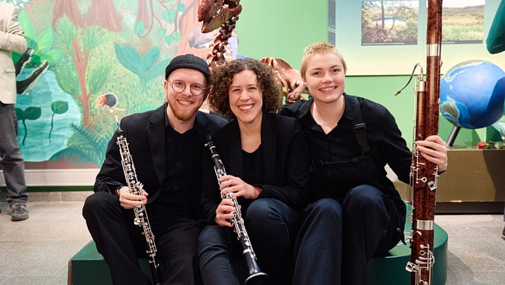 Die Mitglieder:innen des Trio d'Anches heureux sitzen ihren Instrumenten unter der Nachbildung eines Skeletts eines großen Laufvogels und lächeln in die Kamera.