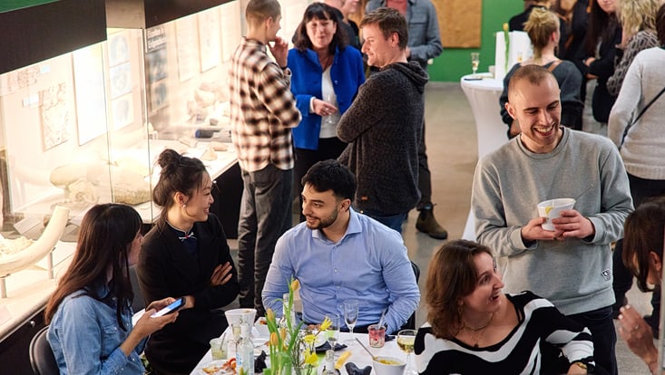 Absolvent:innen sitzen um einen Tisch, vor ihnen Blumen, Teller und Gläser und sie sprechen lächelnd miteinander.